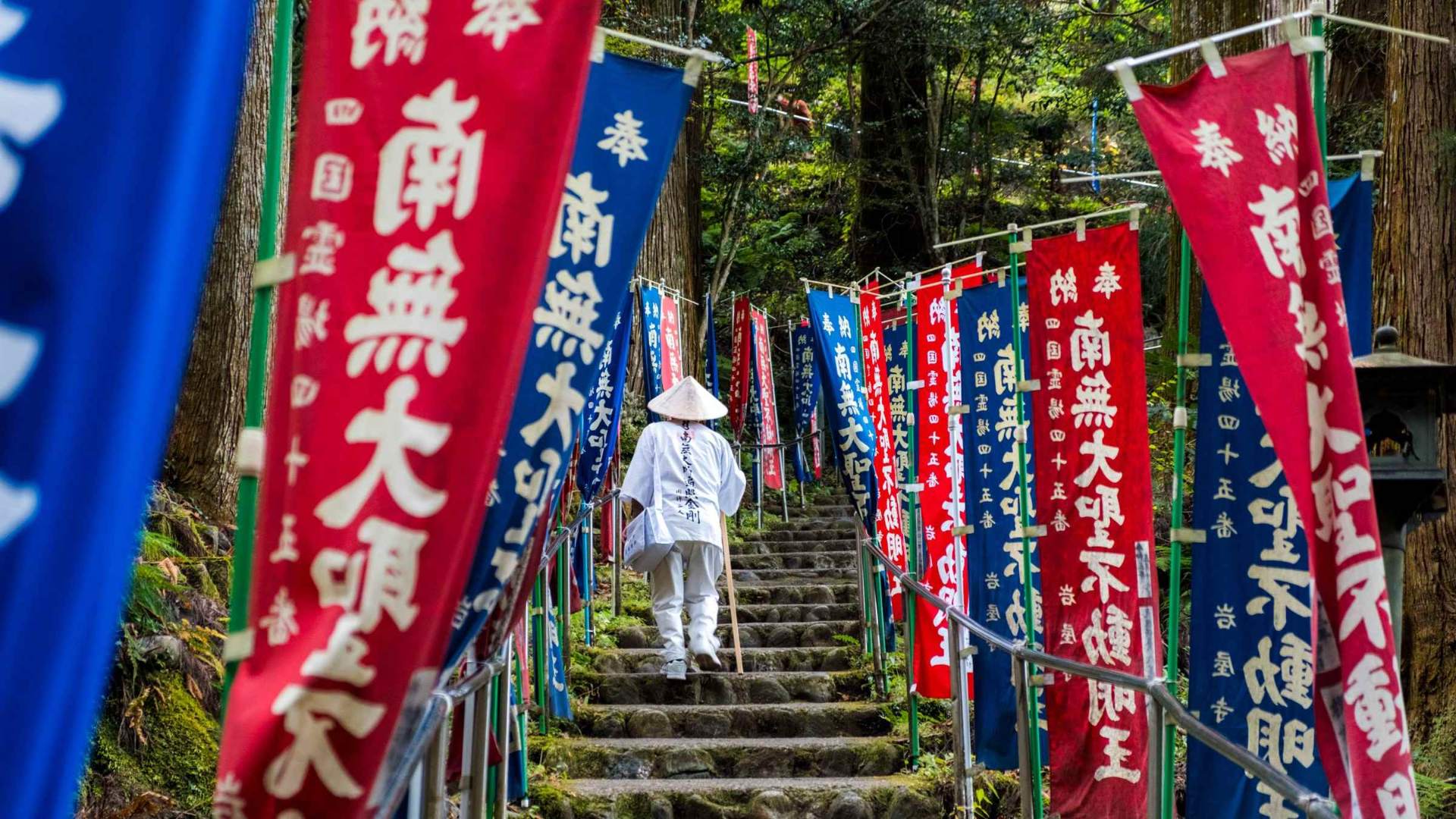 Henro pilgrim ascending stone steps lined with prayer banners at Iwayaji Temple